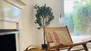 Un salón con chimenea, un sillón de mimbre, una cesta tejida y vistas al verdor a través de un ventanal.