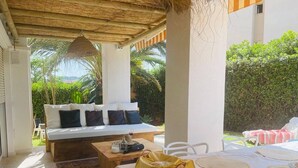 Un espacio al aire libre cubierto con sofá y mesa de comedor, rodeado de follaje.