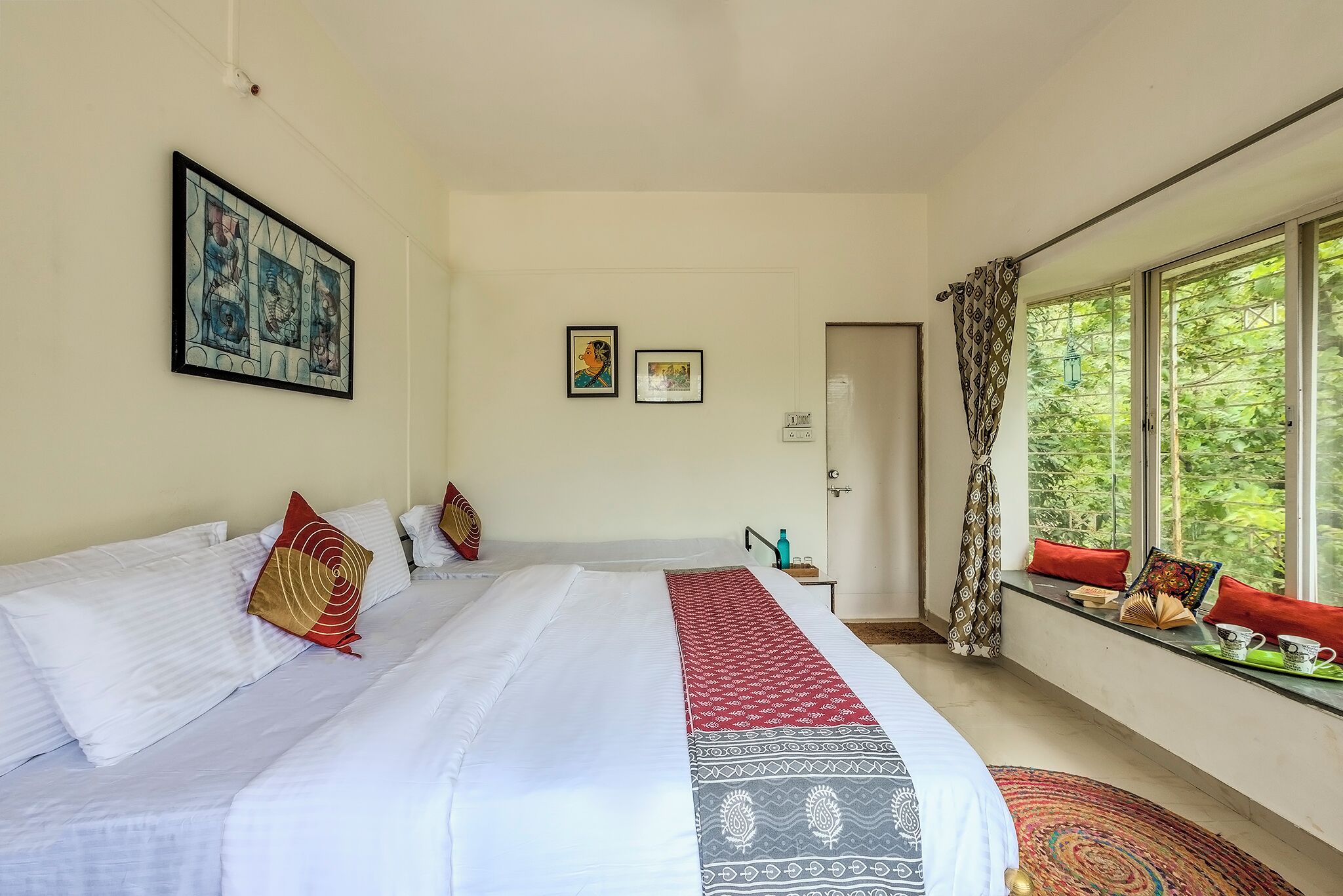 A hotel room with a large bed, a window with a view of greenery, a door, and a small table with a water bottle.