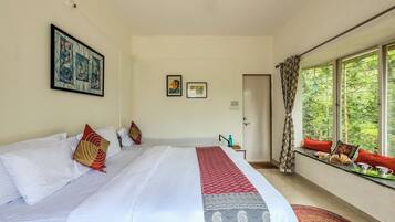 A hotel room with a large bed, a window with a view of greenery, a door, and a small table with a water bottle.