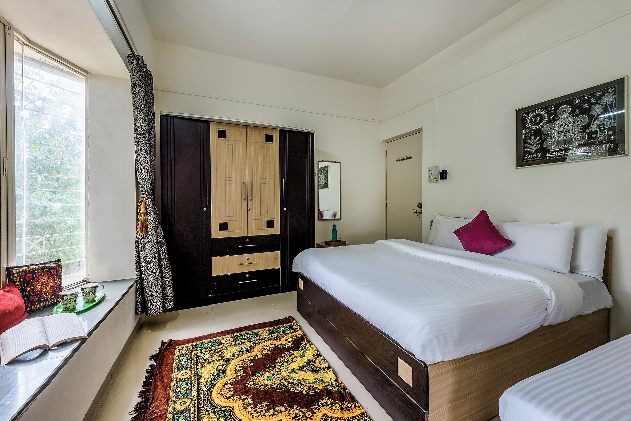 Un dormitorio con un armario grande, una cama con ropa de cama blanca, una alfombra estampada y una ventana con cortinas.