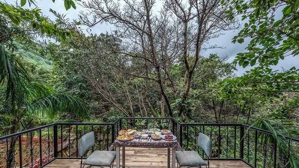 Una terraza de madera con una mesa dispuesta para una comida, rodeada de sillas, y vistas a un bosque frondoso.