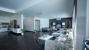 A modern living room with a large sectional sofa, a coffee table, and a TV. There is a kitchen area with cabinets and a countertop visible in the background. - Magnolia - 3BR Private Pool Villa (Kanakapura)