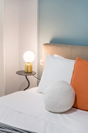 A neatly made bed with white and orange pillows, a round white pillow, and a small table with a lamp beside it.