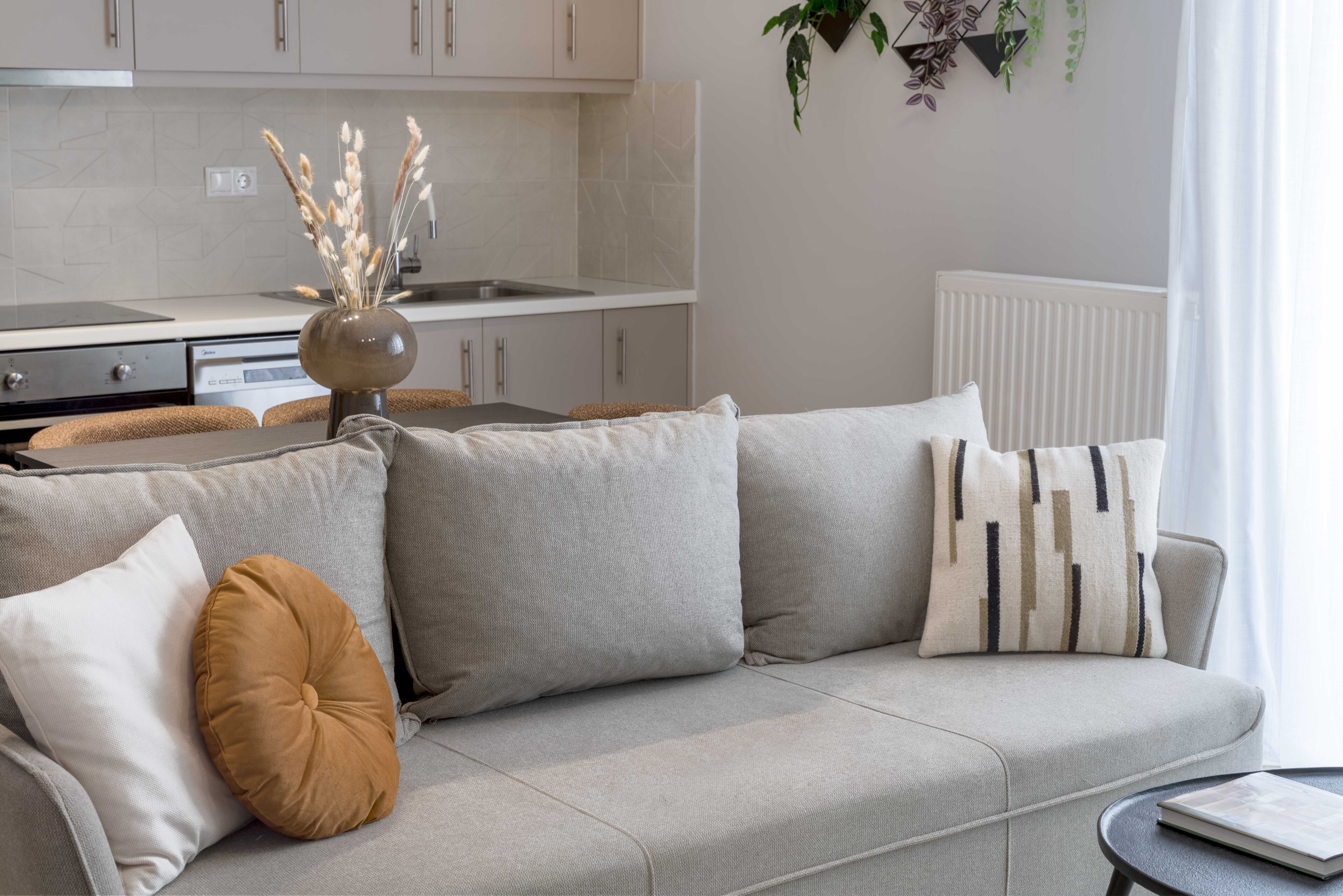 A modern living room with a grey sofa, multiple cushions, a small table with a book, and a kitchen in the background.
