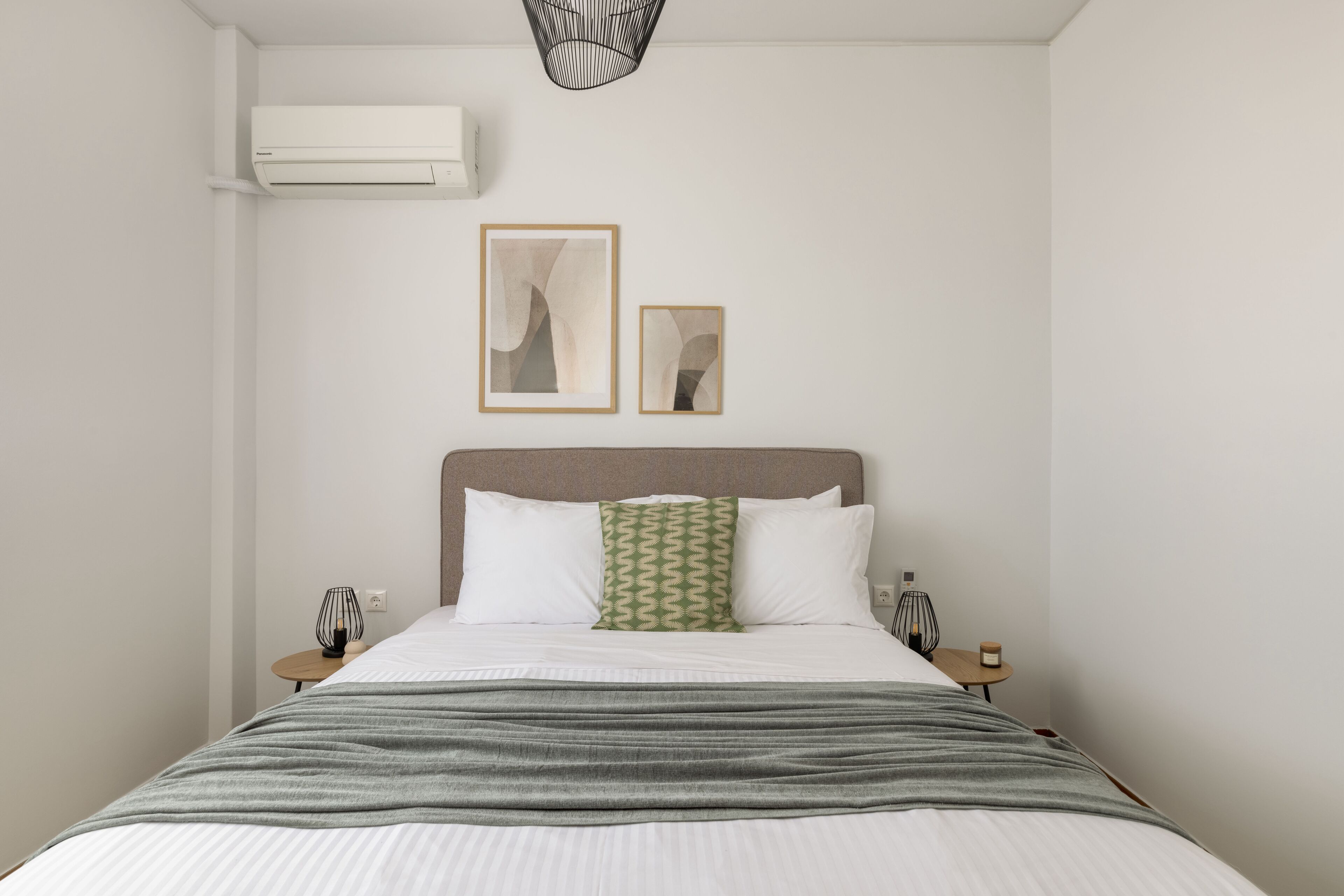 A neatly arranged hotel room with a bed, two bedside tables, a wall-mounted air conditioner, and two framed artworks.