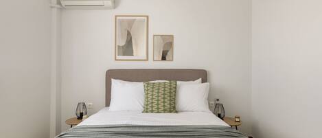 A neatly arranged hotel room with a bed, two bedside tables, a wall-mounted air conditioner, and two framed artworks.