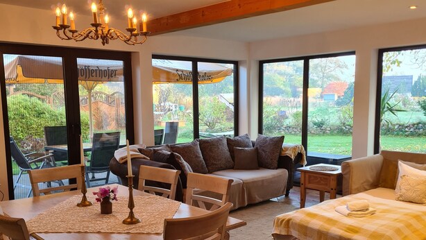 A spacious room with a dining table, chairs, a sofa, and a bed. Large windows offer views of the outdoors. - Romantic house with sauna and fireplace near the lake (Zossen)