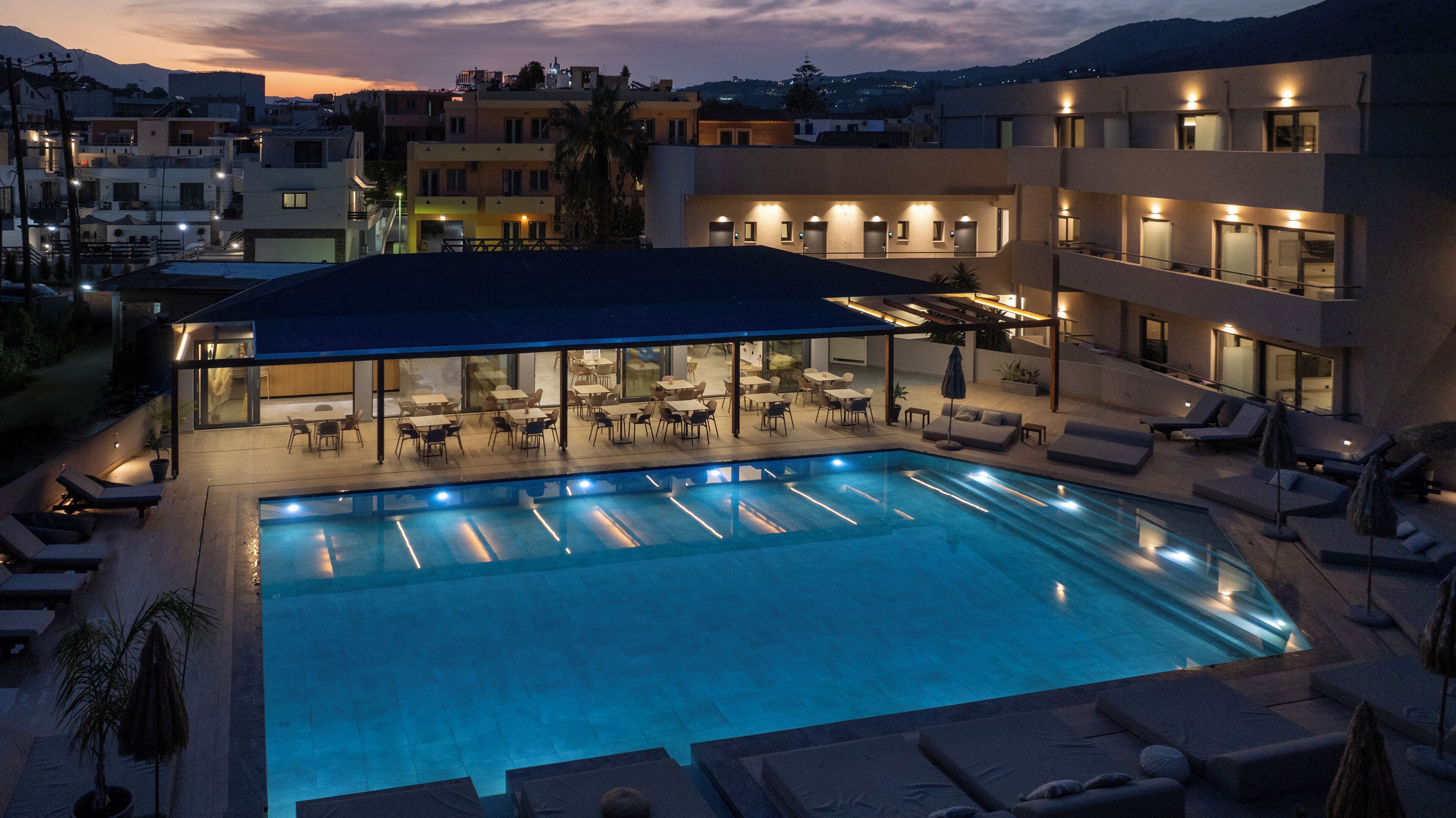 A rooftop pool area with lounge chairs and tables, surrounded by buildings with multiple floors and balconies.