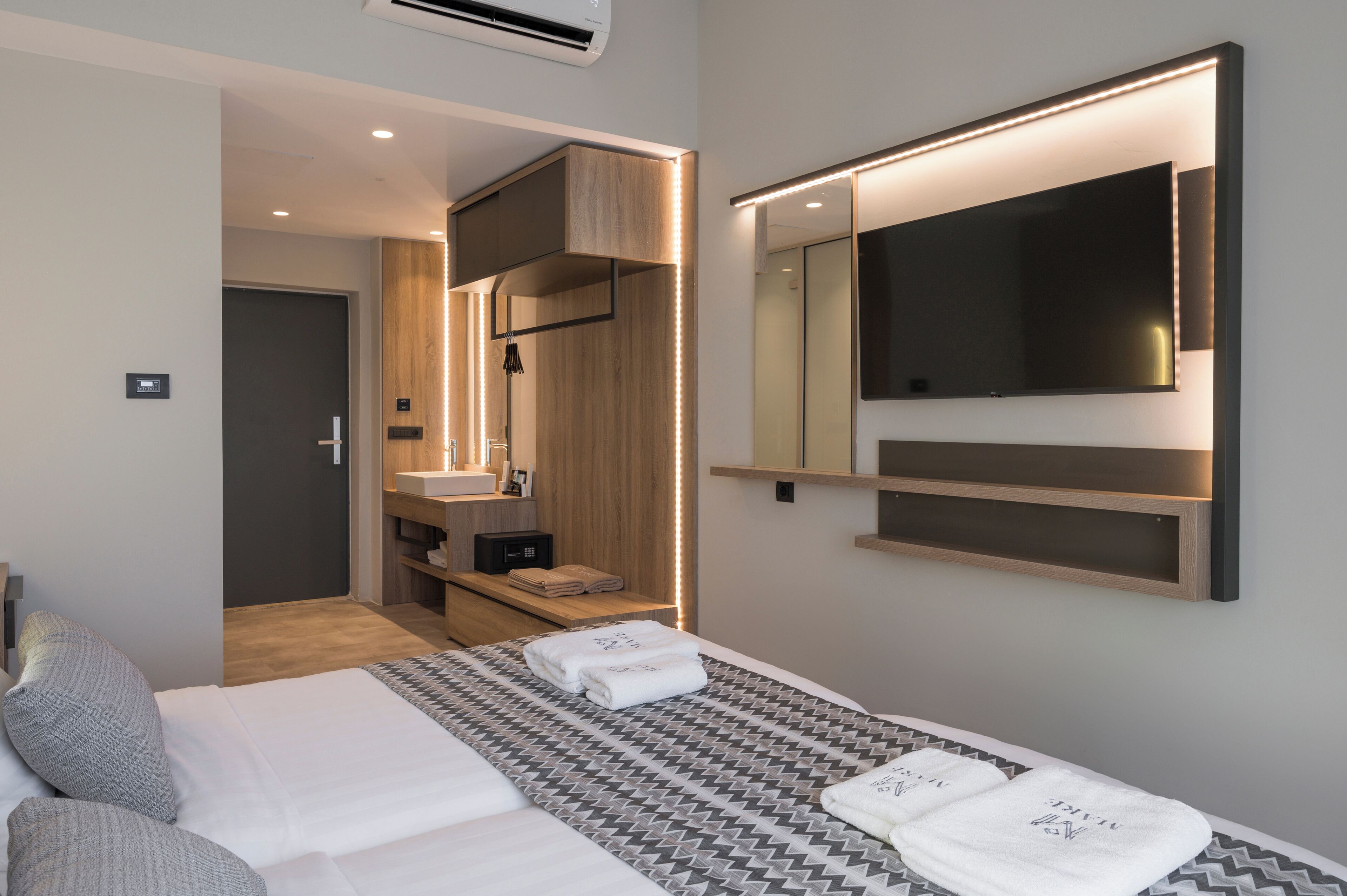 Una habitación de hotel moderna con una cama grande, un televisor montado en la pared, un baño con espejo y lavamanos, y un vestidor.