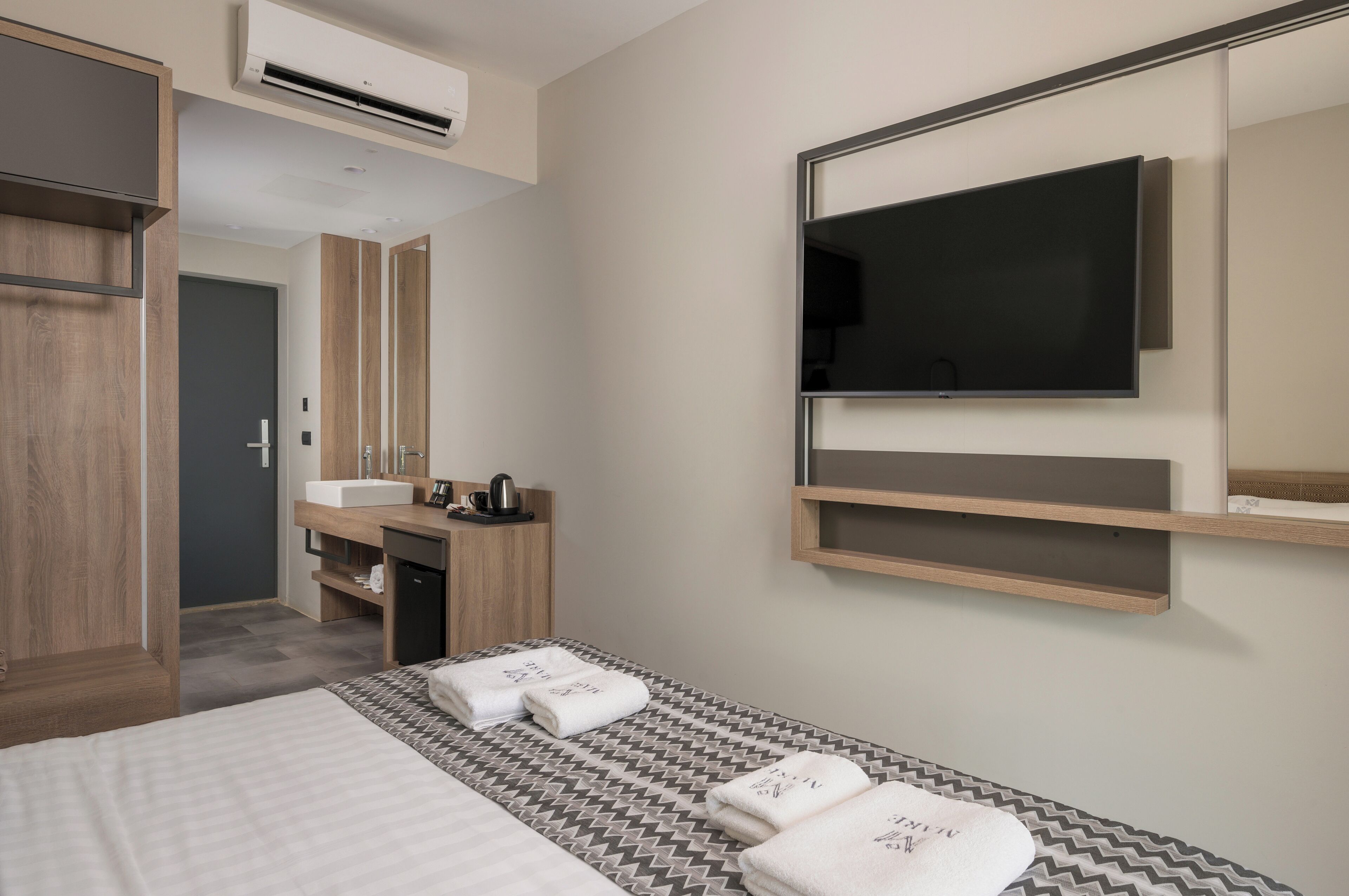 Una habitación de hotel moderna con cama, televisor montado en la pared, armario de madera, baño con lavabo y espejo, y una estantería sobre la cama.