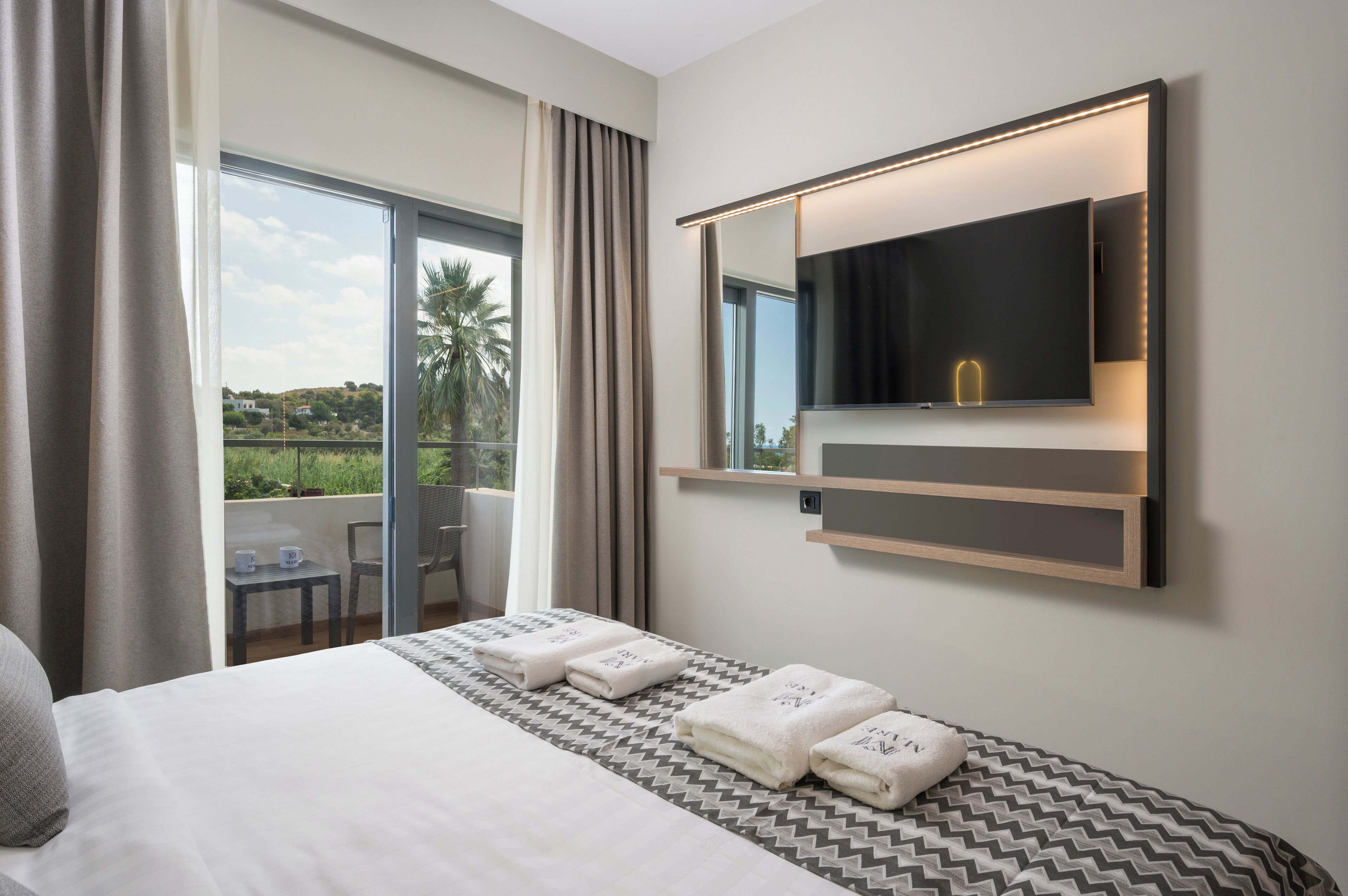 A hotel room with a large bed, a TV mounted on the wall, a balcony with a table and chairs, and a view of greenery outside.