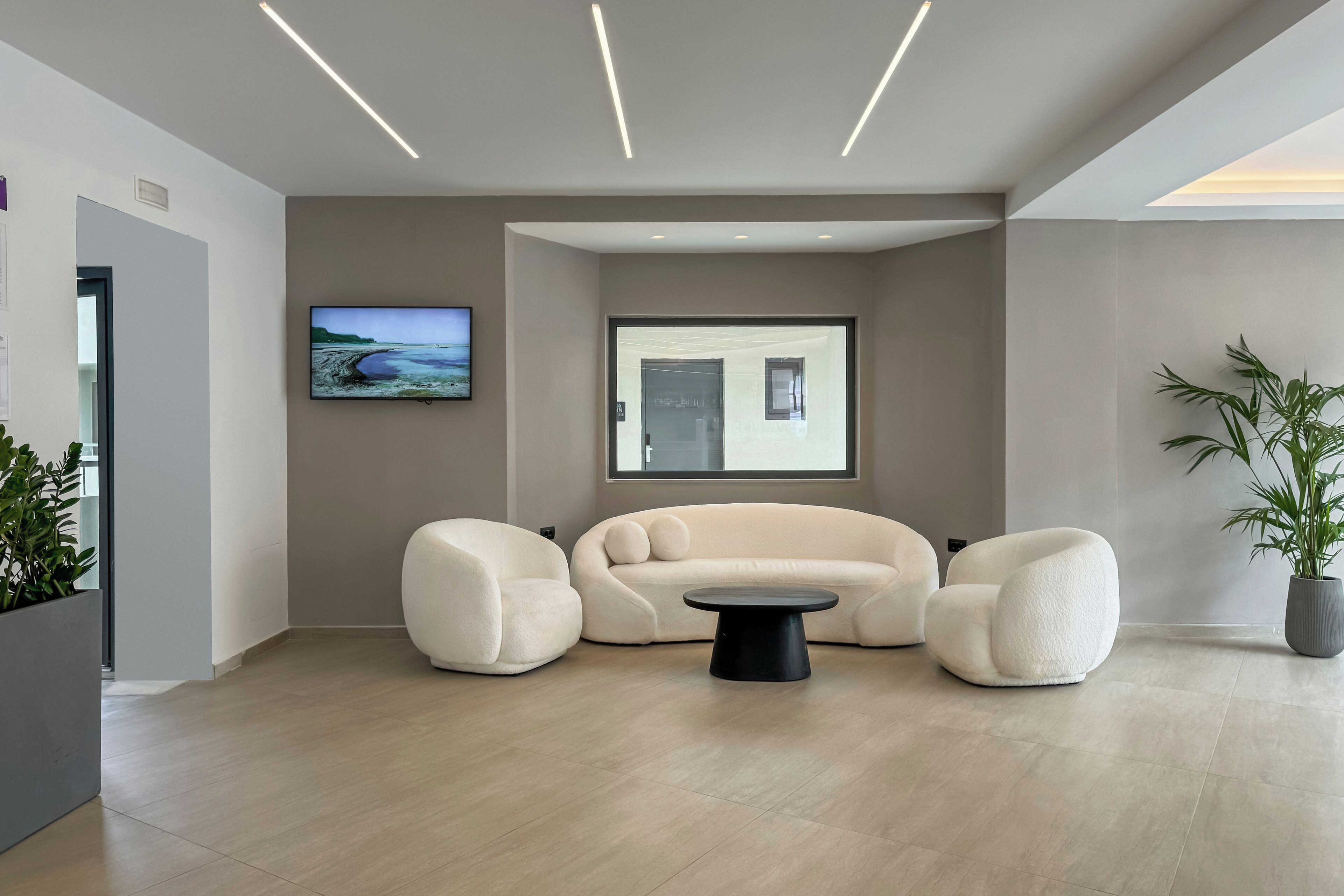 Una sala de estar moderna con un sofá color beige, dos sillones, una mesa de centro negra, un televisor de pantalla plana y un ventanal grande.