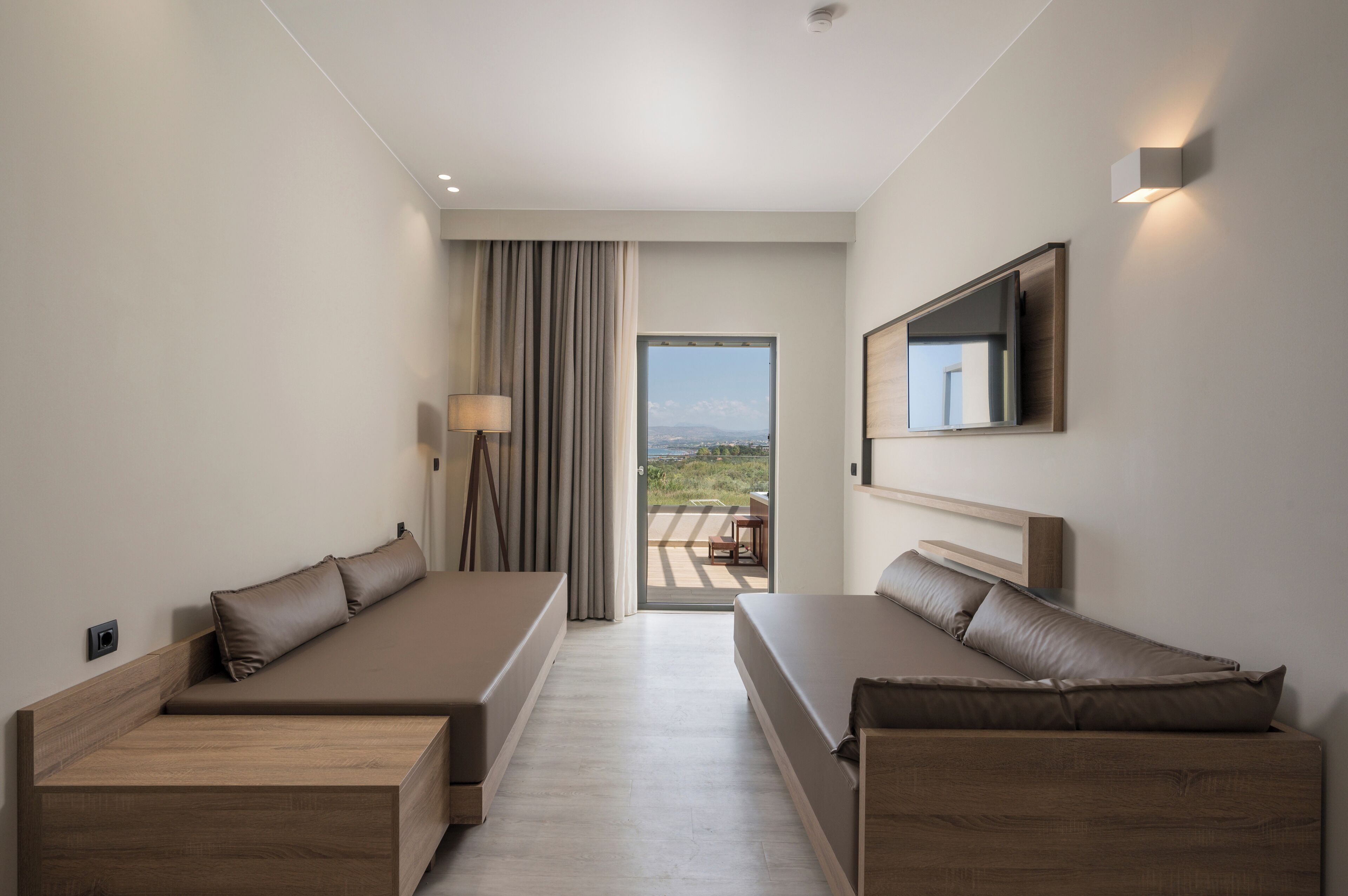 A modern hotel room with a large bed, a sofa, a TV mounted on the wall, and a view of the outdoors.