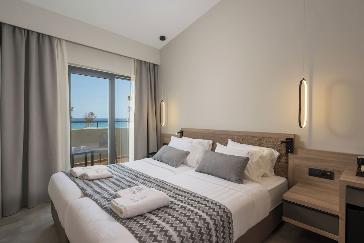 A hotel room with a large bed, a bedside table, a nightstand, and a view of the sea through a sliding glass door.