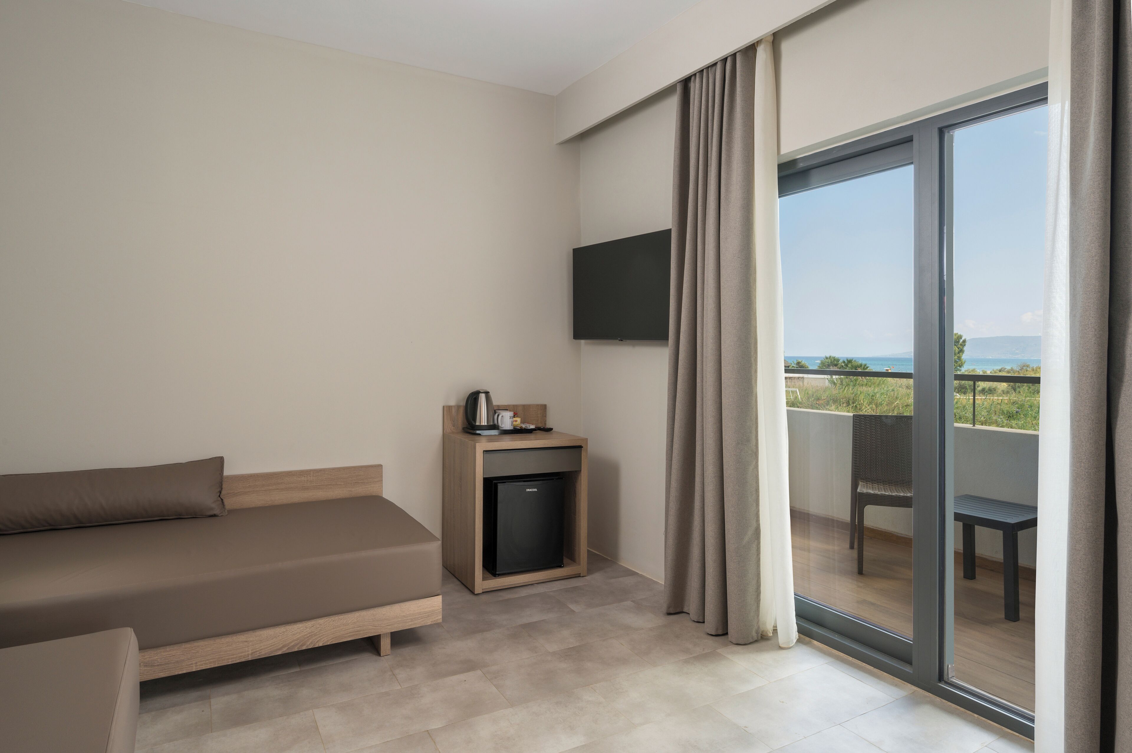 A modern hotel room with a sofa, a small table with a kettle, a flat-screen TV, and a balcony with a view of the sea.