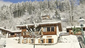 Un edificio cubierto de nieve con fachada de madera, una escalera que sube y un paisaje de árboles nevados al fondo.