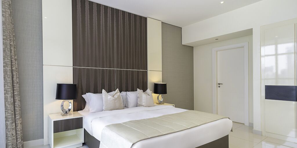 A modern hotel bedroom with a large bed, two bedside tables, a wardrobe, and a large window.