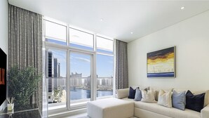 Una sala de estar moderna con un ventanal que ofrece vistas a la ciudad, un sofá color beige con varios cojines y un cuadro enmarcado en la pared.
