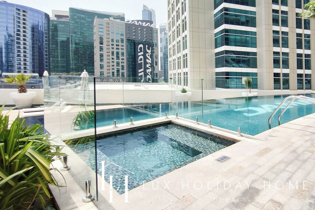 A rooftop pool area with glass railings, surrounded by modern buildings and potted plants.