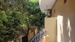 Un étroit couloir extérieur, doté d’une rambarde en métal, d’un mur de couleur beige et d’un arbre qui offre de l’ombre.
