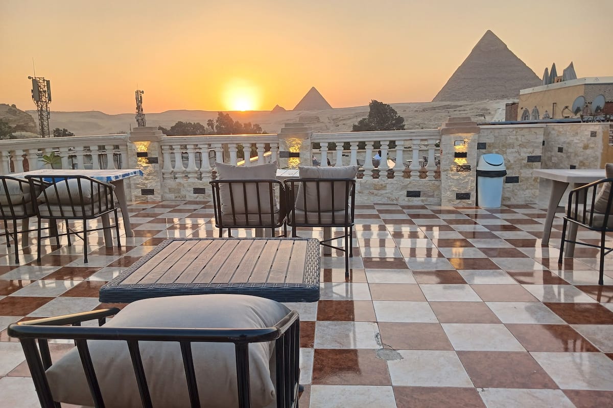 A terrace with checkered flooring, outdoor seating, and a view of the pyramids at sunset.