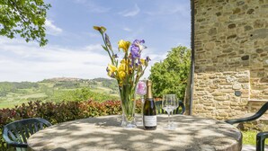 A round stone table with a bottle of wine and glasses, surrounded by chairs, set against a stone wall with a scenic view of hills and a village in the background.