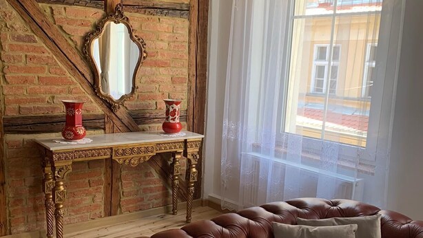 A room with a brown leather sofa, a wooden console table with two red vases, a mirror, and a window with sheer curtains.