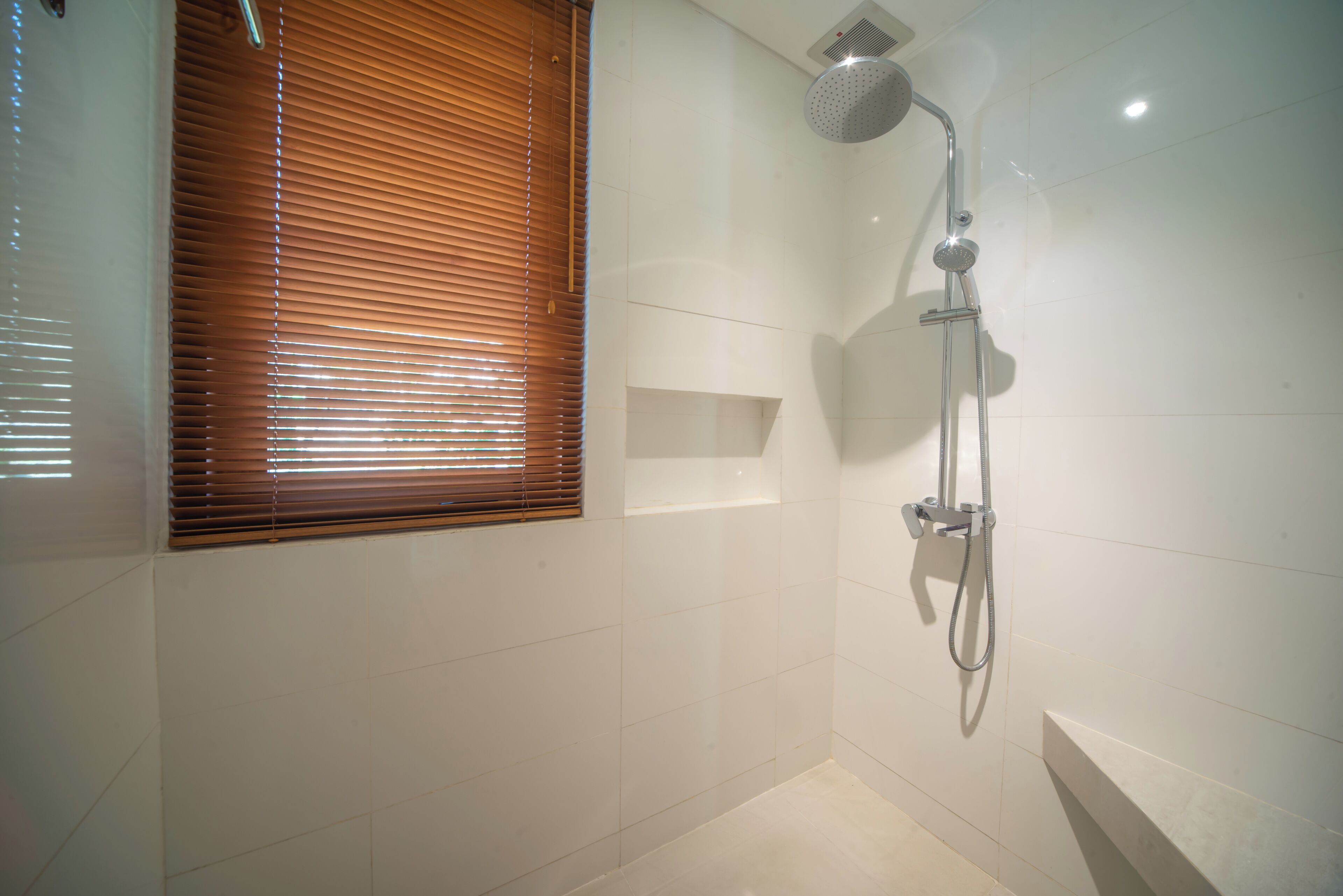 Une salle de bain avec une douche, une fenêtre avec des stores et un mur recouvert de carreaux blancs.