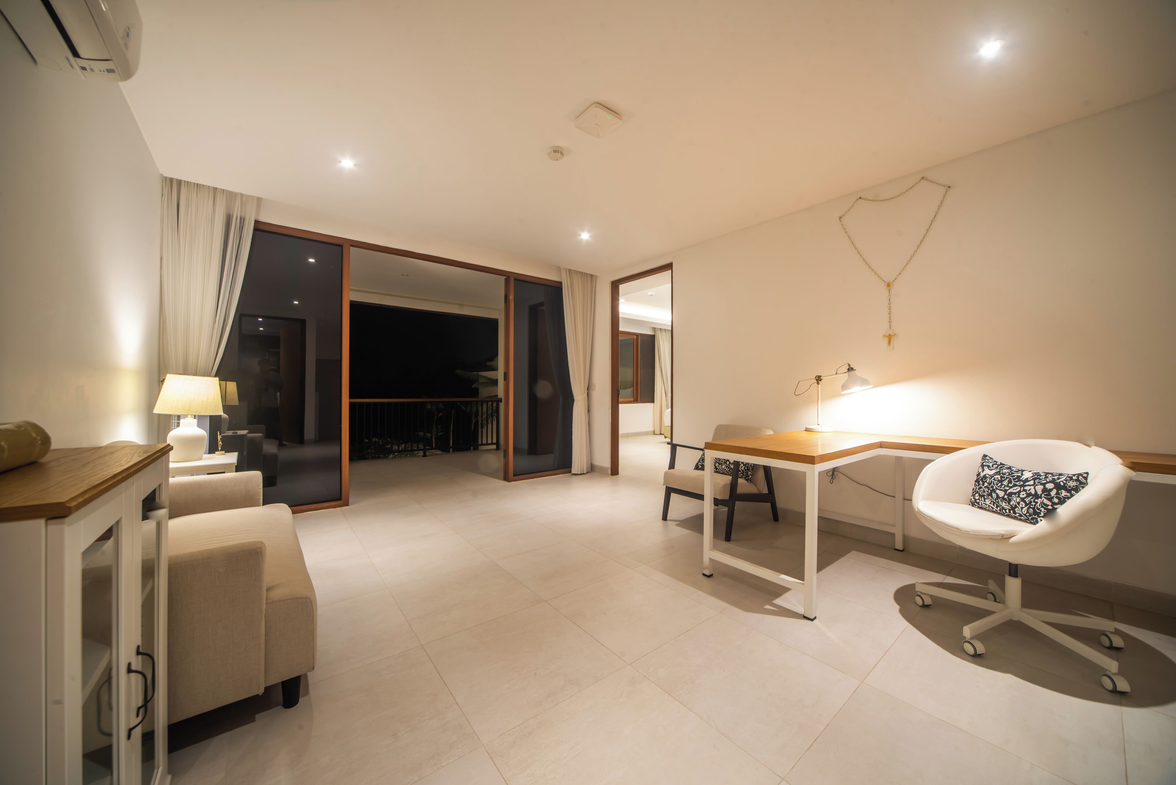 A modern hotel room with a sofa, a desk, a chair, a lamp, and a sliding door leading to a balcony.