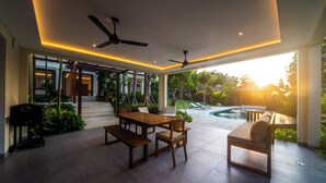 A modern outdoor patio with a wooden table and chairs, ceiling fans, and a view of a swimming pool and garden. - Luxury 4 Bedroom Bella Vista Villa (Ungasan)