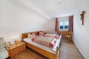 A bedroom with a wooden bed, bedside tables, a chair, a window with curtains, and a crucifix on the wall.
