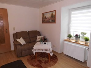 Een woonkamer met een bruine bank, een kleine tafel met een witte tafelkleed, een schilderij aan de muur en een raam met planten op de vensterbank.