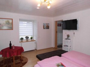 Een kamer met een bed, een tafel met een rode tafelkleed, een raam met jaloezieën, een radiator, een aan de muur bevestigde televisie, en een deur die naar een andere kamer leidt.