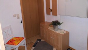 Une petite pièce avec une porte en bois, une chaise blanche avec un coussin à motifs floraux, un meuble en bois orné d’une plante et un miroir accroché au mur.