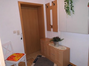 A small room with a wooden door, a white chair with a floral cushion, a wooden cabinet with a plant, and a mirror on the wall. - Apartment 'Komfortable Ferienwohnung' with Private Terrace, Private Garden and Wi-Fi (Gotthun)