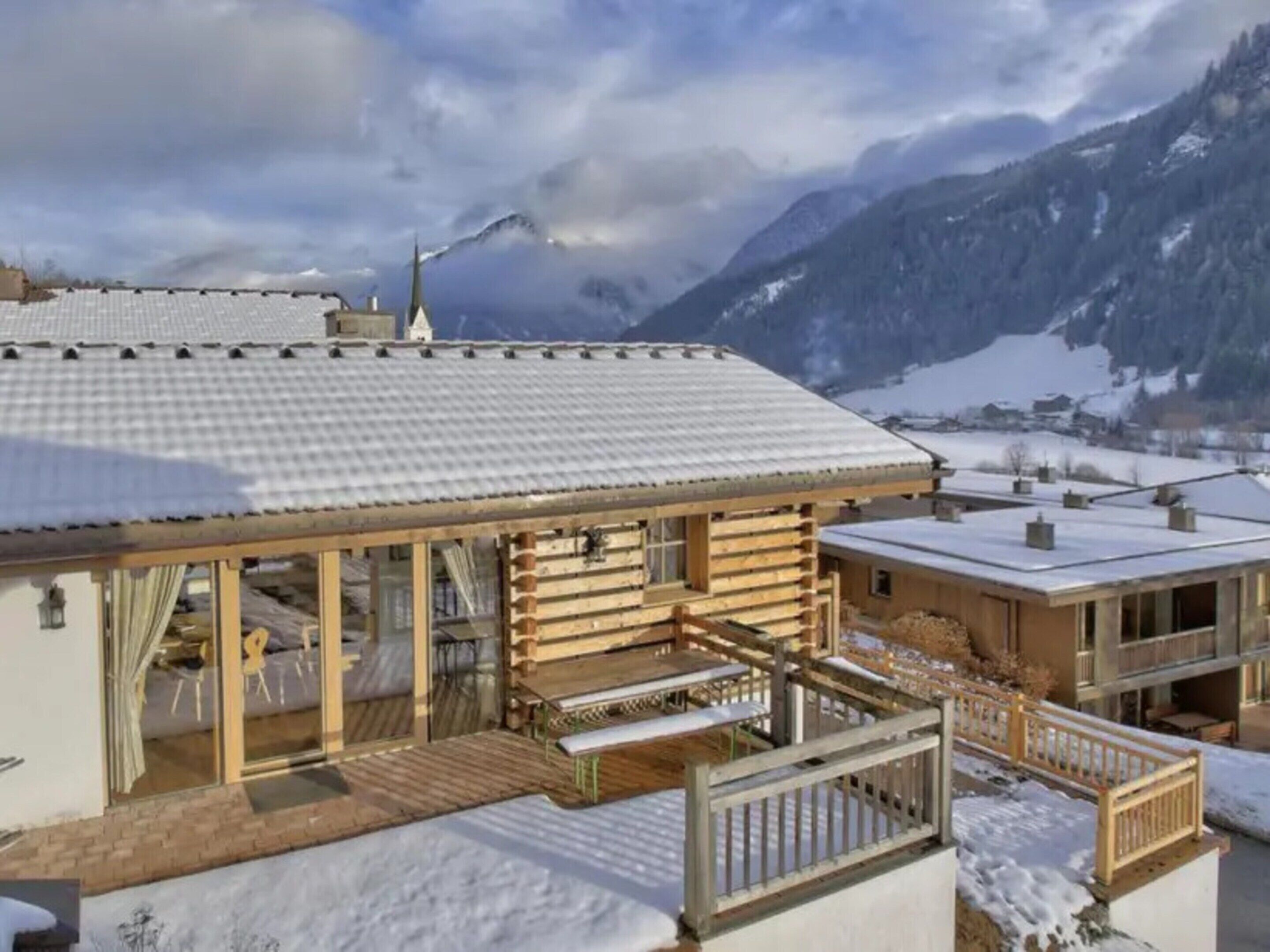 Een houten chalet met balkon, overdekt terras en uitzicht op besneeuwde bergen.