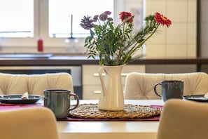 Une table à manger avec un vase blanc contenant des fleurs, une tasse noire et une assiette avec une fourchette.