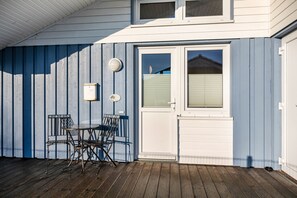 Eine überdachte Terrasse mit Holzdeck, einer weißen Tür mit Fenster sowie einem Metalltisch und Stühlen.