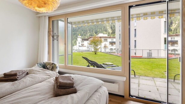 Ein Schlafzimmer mit großem Fenster, einem Bett und Blick auf ein Gebäude sowie Grünfläche.
