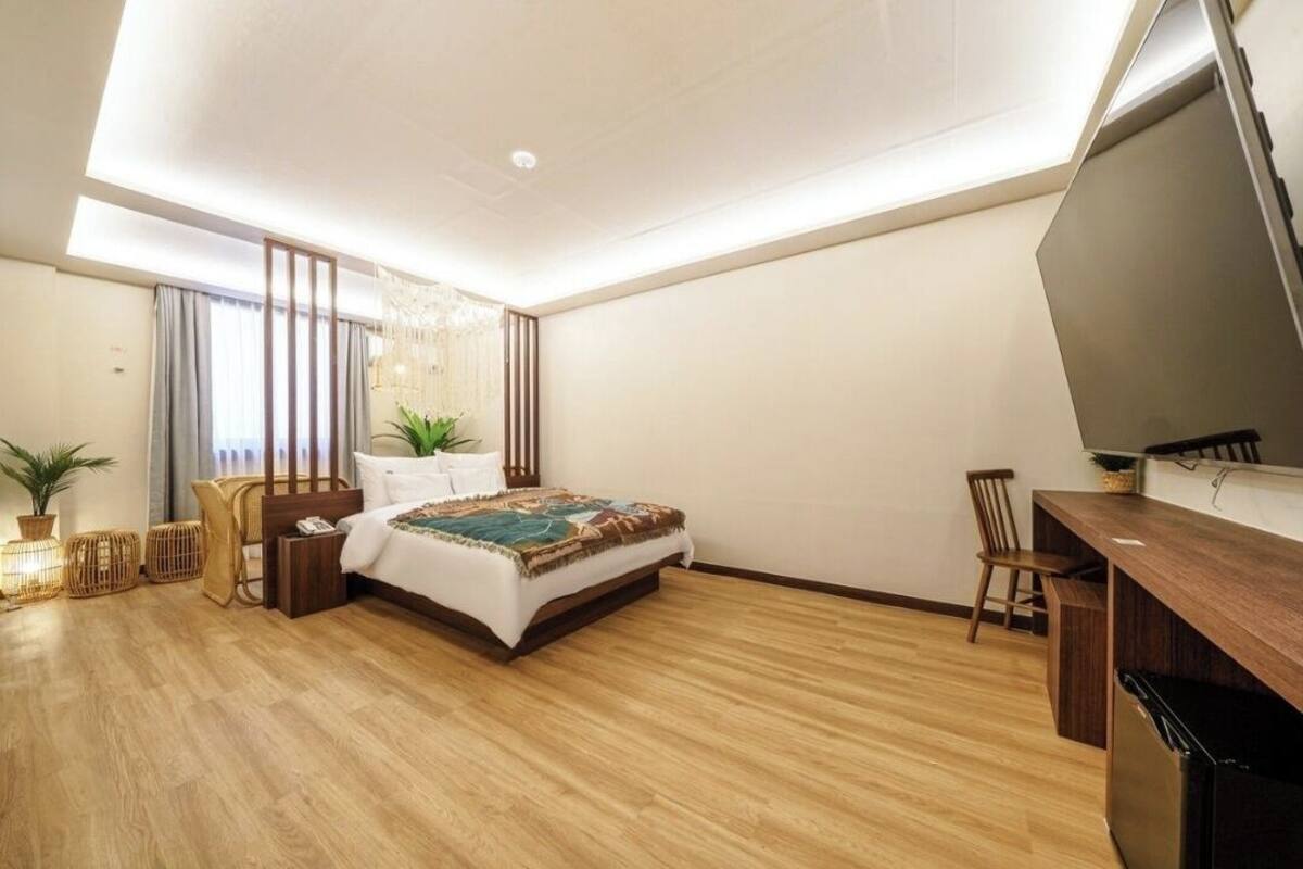 A modern bedroom with a wooden floor, a bed with a colorful blanket, a desk with a chair, and a TV on the wall.
