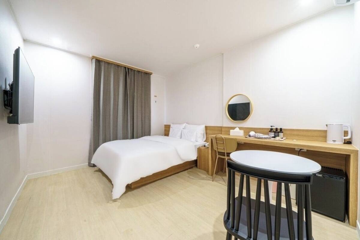 A hotel room with a bed, a desk, a chair, a round table, a small refrigerator, and a television.