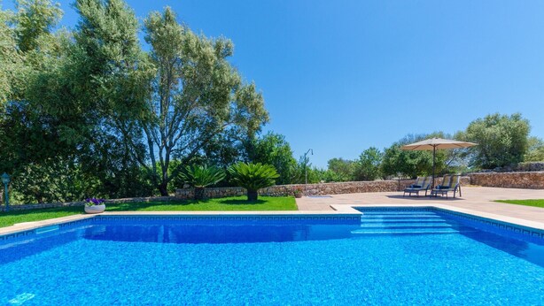 Piscina azul cristalina con sillas de descanso y una sombrilla, rodeada de árboles y un muro de piedra.