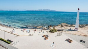 Ein Strand mit einem hohen weißen Leuchtturm, klarem blauen Meer, felsiger Küstenlinie und Menschen, die die Sonne genießen.