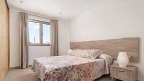A bedroom with a bed, a nightstand, a window with curtains, and a view of the city. - Ca Na Xisca 2 - Apartment In Can Picafort (Can Picafort, Illes Balears)