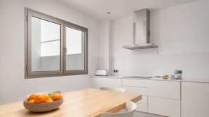 A modern kitchen with white cabinetry, a wooden dining table, and a bowl of oranges on the table. - Ca Na Xisca 2 - Apartment In Can Picafort (Can Picafort, Illes Balears)