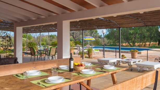 Un espacio de comedor al aire libre con techo, con una larga mesa de madera dispuesta para una comida, rodeada de sillas y una piscina al fondo.