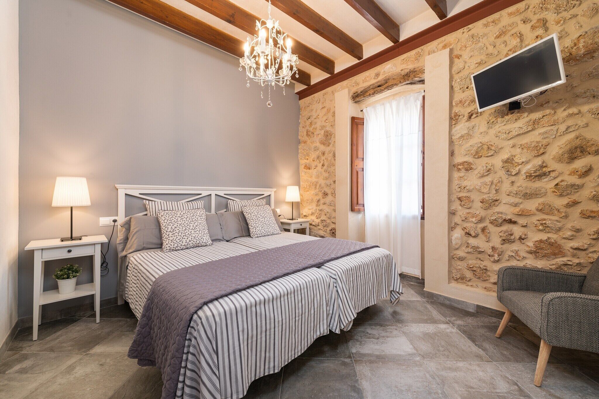 A bedroom with a bed, bedside lamps, a TV, a chandelier, a chair, and a stone wall.
