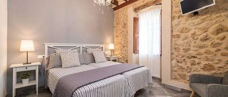 A bedroom with a bed, bedside lamps, a TV, a chandelier, a chair, and a stone wall.