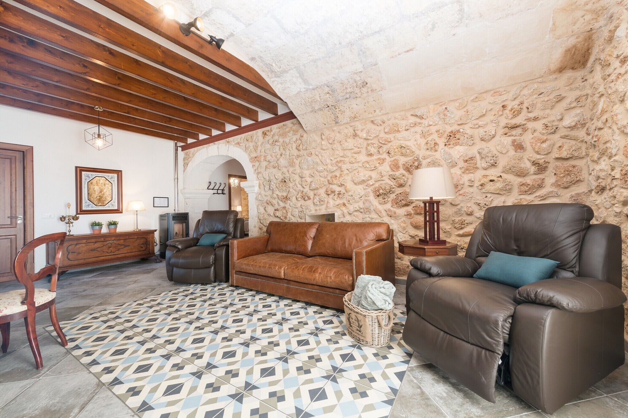 A cozy living room with a stone wall, wooden ceiling beams, a patterned rug, leather sofas, and a wooden cabinet.